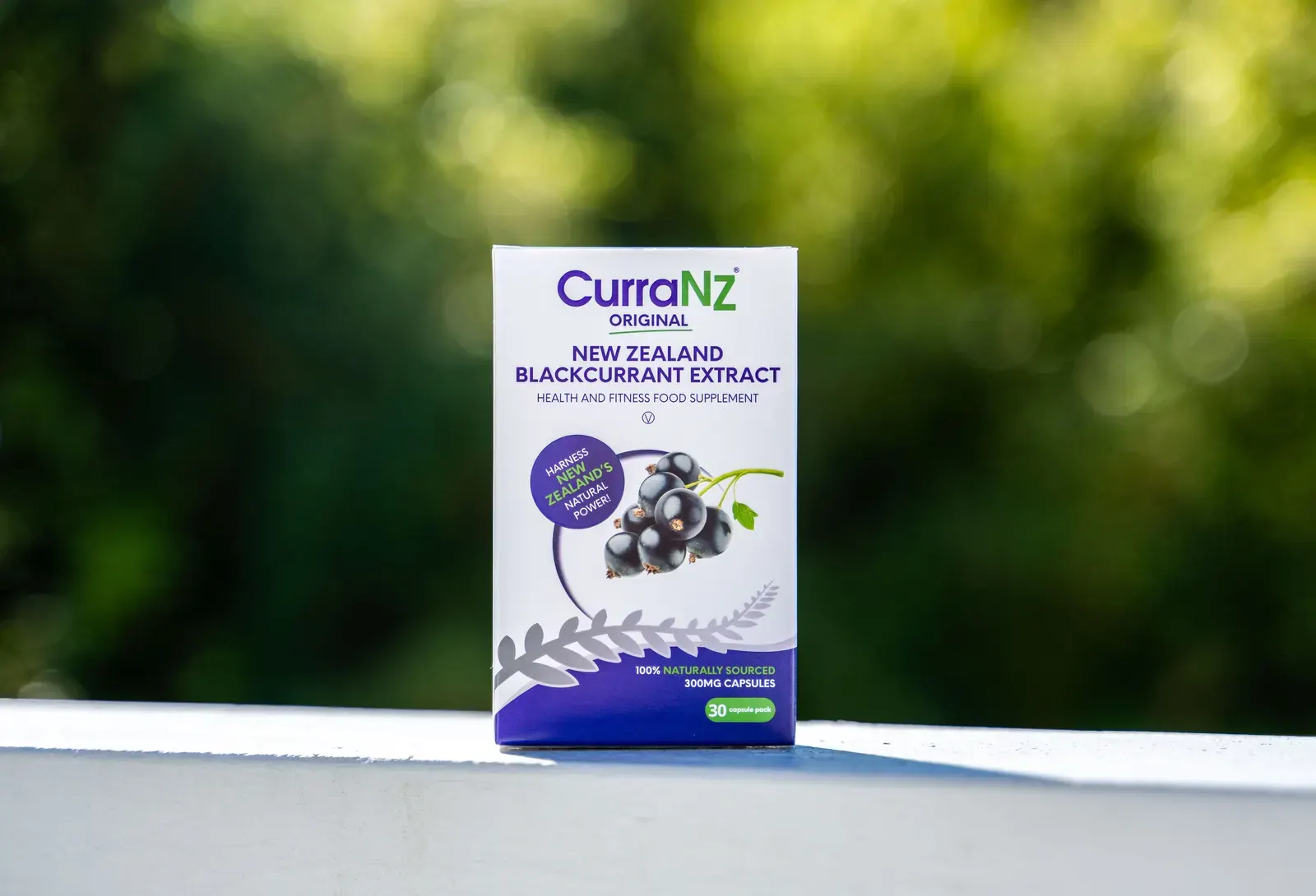 A box of CurraNZ New Zealand blackcurrant extract capsules on a white ledge against a blurred green garden.