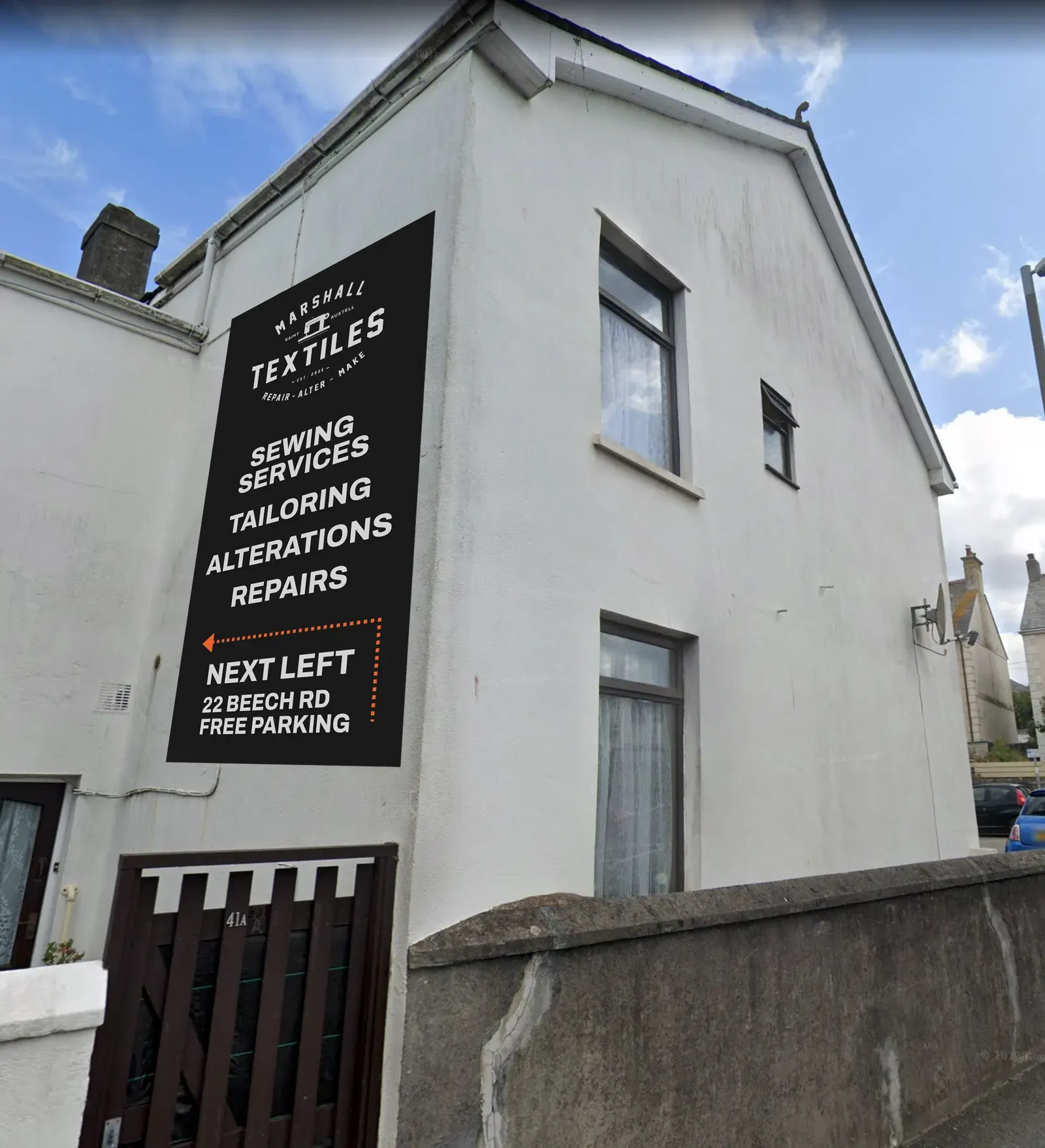 A white building in St Austell with a tall black Marshall Textiles sign listing sewing services, tailoring, alterations and repairs.