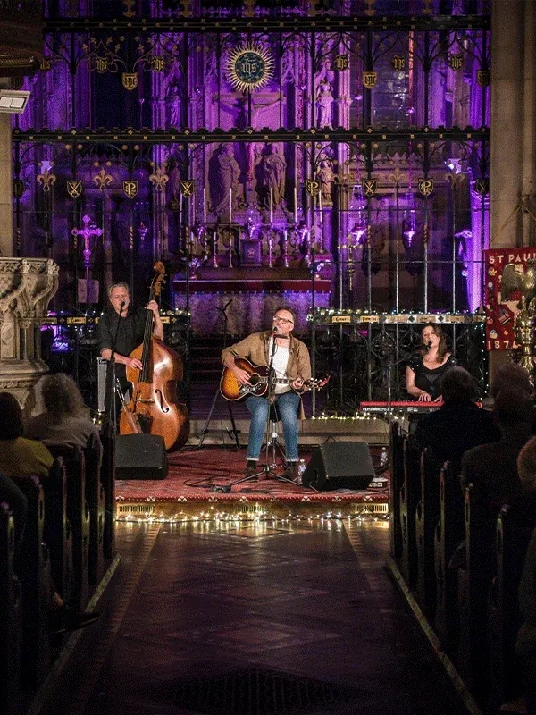 An Aftershocks acoustic gig inside a candlelit church lit in purple, with a double bass, a guitarist and a keyboard player at the altar steps.