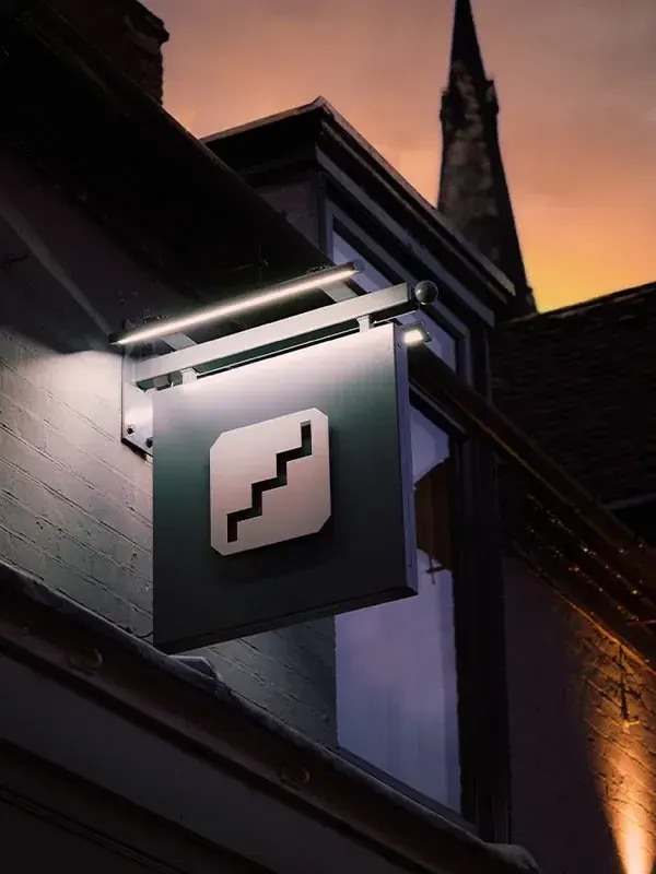 The Upstairs hanging sign, a white stair mark on a slate panel, lit from above against a dusk sky with a church spire behind.